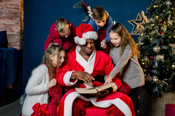 Op kerstavond leest Santa Claus het kerstverhaal voor kinderen die naar hem van alle kanten, op de achtergrond van een kerstboom luisteren. — Stockfoto