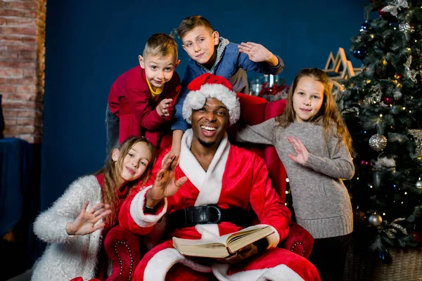 Santa Claus lee un libro antiguo con cuentos de Navidad para niños sentados alrededor de una silla roja — Foto de Stock