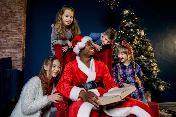 Afrikaanse kerstman leest kinderen kerstverhalen in een gezellige ingerichte Oud en Nieuw kamer. — Stockfoto