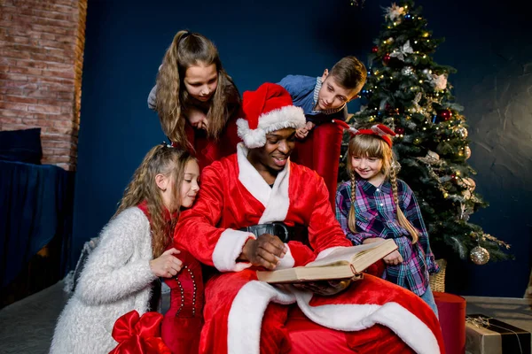 Babbo Natale legge un antico libro con storie di Natale per bambini seduti intorno a una sedia rossa — Foto Stock