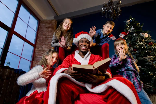 En Nochebuena, Santa Claus lee el cuento de Navidad para los niños que lo escuchan desde todos los lados, en el fondo de un árbol de Navidad . — Foto de Stock