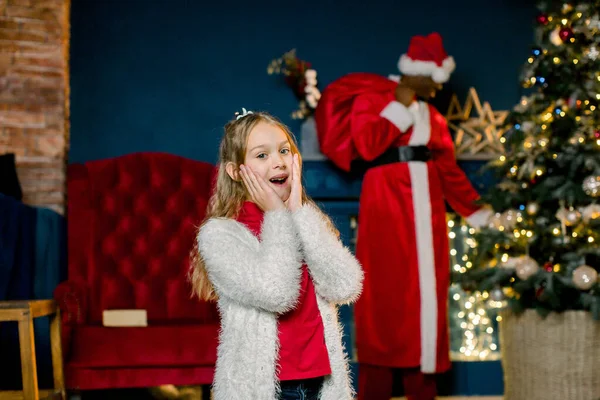 Carino bambina è felice ed eccitato in piedi in accogliente stanza decorata. Babbo Natale africano è venuto e mette regali all'albero di Natale . — Foto Stock