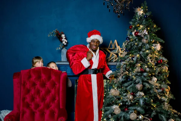 Babbo Natale africano mettere regali sotto l'albero di Natale in accogliente stanza decorata, mentre due bambine eccitate guardano fuori dalla sedia rossa — Foto Stock