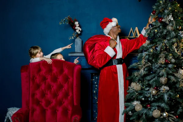 Afrikaanse Kerstman zet giften onder een kerstboom, en ziet niet dat twee kleine meisjes niet slapen en gelukkig voor hem kijken — Stockfoto