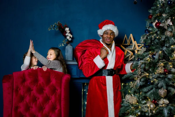 Babbo Natale africano mette i regali sotto un albero di Natale, e non vede che due bambine non dormono e sembrano felici per lui — Foto Stock