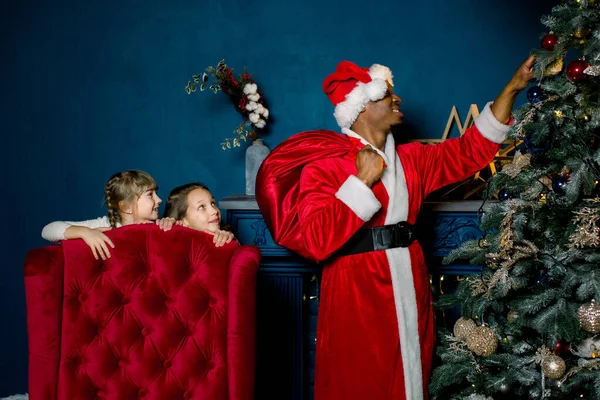 Kerstman zet geschenken onder kerstboom in gezellige blauwe kamer. Afrikaanse kerstman zet geschenken onder een kerstboom, en twee kleine meisjes slapen niet en kijken blij voor hem — Stockfoto