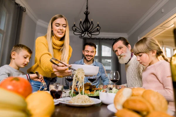 Glücklich lächelnde Familie, Großvater, Eltern und Kinder sitzen am festlichen Tisch und essen leckeres Abendessen. Mutter legt Nudelspaghetti auf Teller für alle Familienmitglieder — Stockfoto