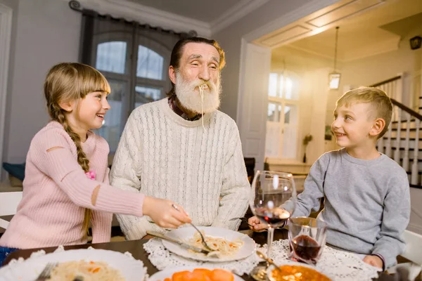 Old handsome grandfather with his two grandchildren sitting at the kitchen table and eating pasta. Little girl and boy feeding grandfather with pasta and laughing. Happy lifestyle family moments.