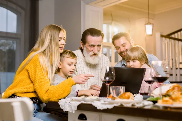 Šťastně se usmívající rodina vypadá film nebo volat přes internet pomocí notebooku, sedí na slavnostní tabe doma, slaví večeři společně. Koncept Dne díkůvzdání — Stock fotografie