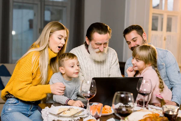 Happy smiling family looking film or making call via internet using laptop, sitting at the festive tabe at home, celebrating dinner together. Thanksgiving day concept — Stock Photo, Image
