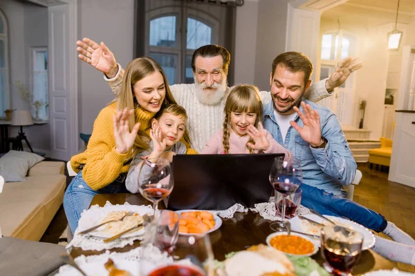 Krásná rodina, sedí u stolu s večeří, usmívá se, je šťastná a používá notebook. Family making videocall using laptop celebrating at home — Stock fotografie