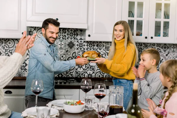 Celebration, holidays and people concept - happy young parents, woman and man, offering roast chicken to family, children and grandfather, having dinner party at home — Stock Photo, Image