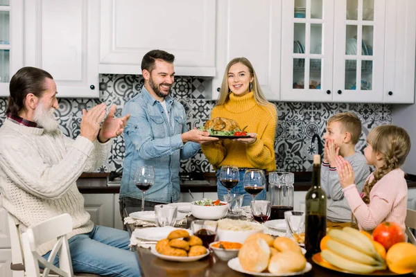 Šťastný Den díkůvzdání. Rodina sedí u stolu a slaví svátky. Tradiční večeře. Tři generace rodin. Mladí rodiče drží pečeného krocana a usmívají se — Stock fotografie