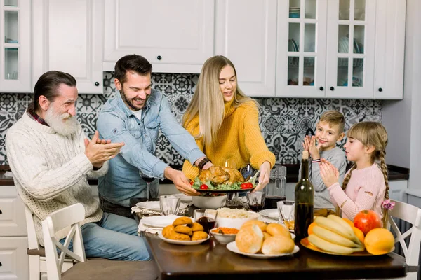 Happy Thanksgiving Day. Family sitting at the table and celebrating holiday. Traditional dinner. Three generations of family. Young parents holding roast turkey and smiling