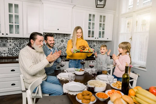 Thanksgiving Dinner concept: Woman Holding Platter With Roast Turkey And Garnish for family dinner at home. All family members are sitting at the table, excited, happy and applaud