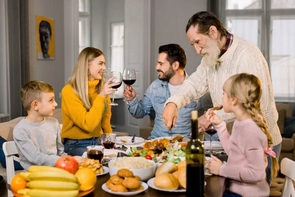 Çocuklarla dolu büyük bir aile, yaşlı büyükbaba hindi keserken ve genç ebeveynler şarap kadehlerini tokuştururken evde lezzetli bir şükran günü yemeği yiyorlar. — Stok fotoğraf