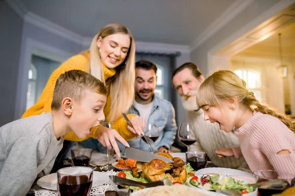 感恩节庆祝传统家庭晚餐的概念。 妈妈正在切火鸡，两个兴奋的小孩张大嘴看着食物，照片上是一家人坐在桌旁的滑稽照片 — 图库照片