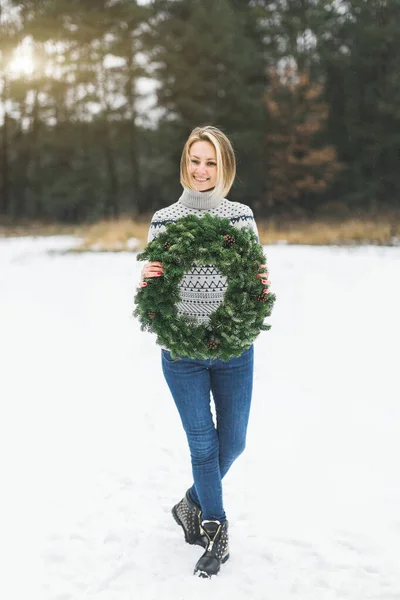 Jovem loira de cabelo curto senhora em camisola de malha cinza mantém grinalda de Natal com neve floresta de inverno no fundo — Fotografia de Stock