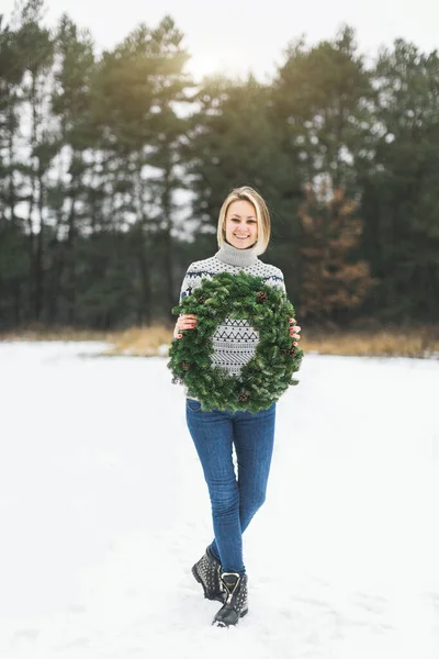 Молода блондинка коротке волосся леді в сірому в'язаному светрі тримає різдвяний вінок зі сніжним зимовим лісом на фоні — стокове фото