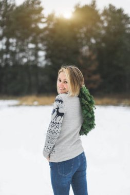 Kış ormanlarında yaşayan bir kızın elinde şık bir Noel çelengi. Kadının arka planı, kameraya bakıp gülümseyen, elinde yeni yıl çelengi tutan.