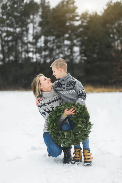 圣诞快乐，节日快乐。 妈妈和她的小儿子一起在冬天的森林里散步，抱着圣诞花环。 他们彼此凝视着，面带微笑. — 图库照片