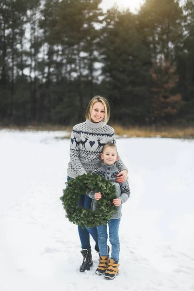 Happy mother with her little son standing on the background of winter forest outdoors and holding Christmas pine wreath — Stock Photo, Image
