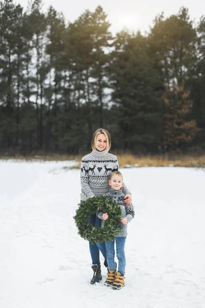 Happy mother with her little son standing on the background of winter forest outdoors and holding Christmas pine wreath — Stock Photo, Image