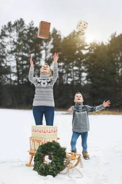 Winter fairy tale, a young mother and her son in the forest. Mom and child throwing gifts on the eve of the new year in the winter forest. Wooden sledge with Christmas wreath and presents on the snow — Stock Photo, Image