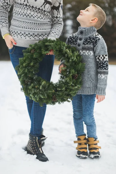 Image recadrée de jambes de mère et de fils, tenant ensemble une belle couronne de Noël, debout à l'extérieur dans un parc d'hiver ou une forêt. Mère et enfant avec couronne de Noël, à l'extérieur — Photo