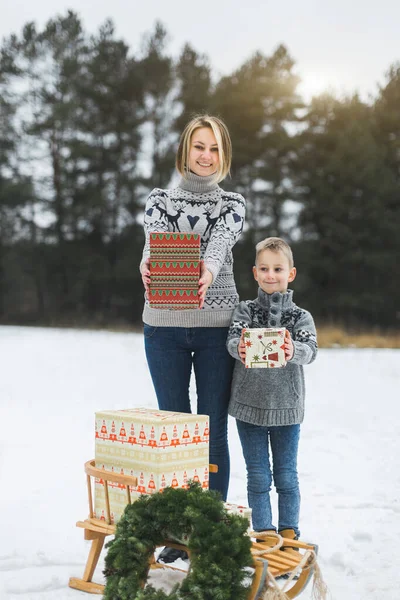 Merry Christmas and Happy Holidays concept. Mother and child son with christmas presents on winter snowy forest background. Portrait of beautiful family standing near the wooden decorated sledge. — Stock Photo, Image