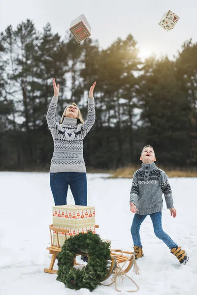 Mutter und Sohn haben Spaß zusammen, springen und werfen Weihnachtsgeschenkboxen hoch. Winterurlaub, Familienwanderung im Winterwald. dekorierte Holzschlitten mit Geschenkboxen — Stockfoto