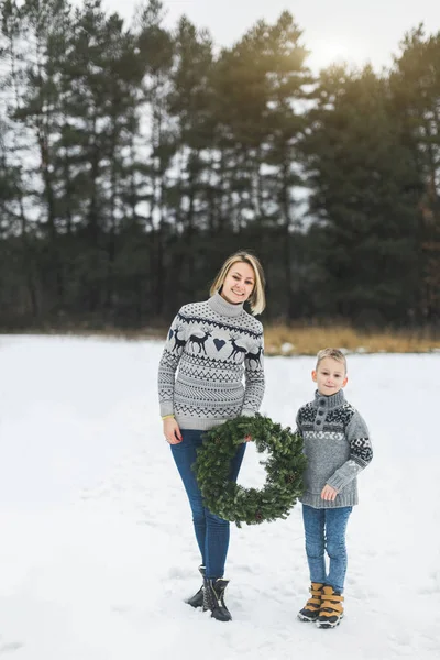 New Year and Christmas Holidays. Caucasian mom with her son holding decorated Christmas wreath in arms while standing outdoors on the background of winter snowy forest. — Stock Photo, Image