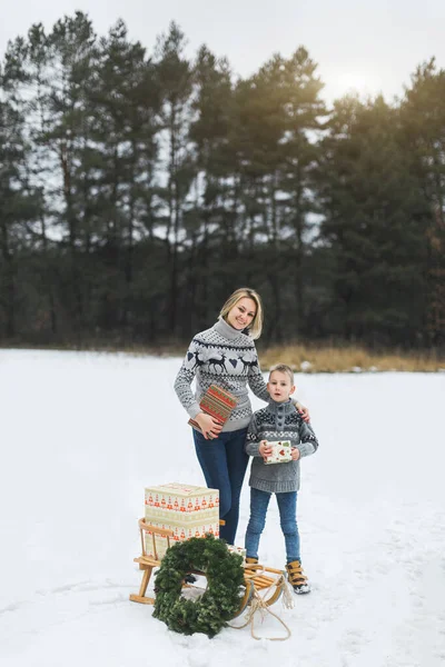 Happy family, mother and her little son, with Christmas gift box in winter forest. Stylish wooden sledge with presents and Christmas wreath on the snow — Stock Photo, Image