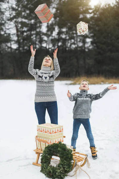 Family, christmas, x-mas, winter, happiness and people concept - smiling family, mom and son, in knitted sweaters, throwing up gift boxes while standing on the background of winter forest — Stock Photo, Image