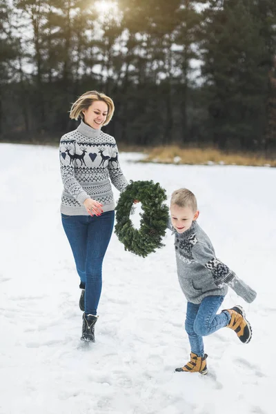 Happy family mother and child son in jeans and knitted sweaters on a winter walk outdoors, running and having fun, holding home made Christmas wreath — Stock Photo, Image