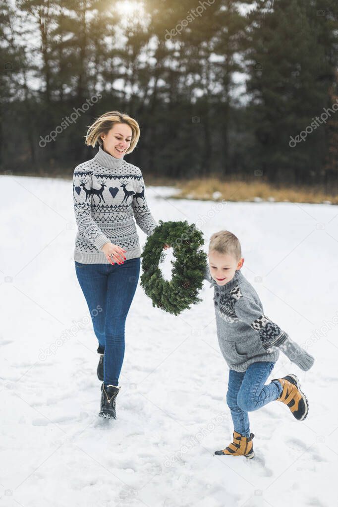 happy family mother and child son in jeans and knitted sweaters on a winter walk outdoors, running and having fun, holding home made Christmas wreath