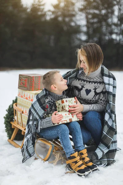 pretty young mother talk with child son while sitting on the wooden sledge outdoors in winter forest, covered with checkered plaid together. Family winter holidays outdoors.