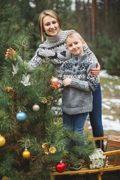 Family, young mother and little son, wearing knitted sweaters, decorating fir tree in the forest or park. Colorful decorations, wooden sledge, Christmas presents — Stock Photo, Image