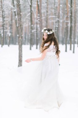 Genç ve güzel bir kadının kışın ormanda yürüyüşünün arka görüntüsü. Kafasında çelenk olan, uzun kıvırcık saçlı, elinde buket tutan gelin.