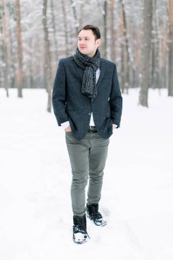 Yakışıklı hippi damat şık bir takım elbise giyiyor, karlı ormanda duruyor, uzun bir fotoğraf.