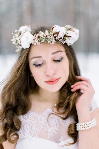 Belle mariée touchant ses cheveux bouclés, avec couronne de bandeau floral, faite de fleurs et de coton, debout à l'extérieur, sur le fond de la forêt d'hiver — Photo