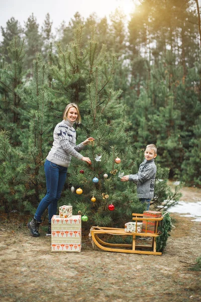 Mother and little son looking at camera and smiling, while decorate Christmas tree in winter forest, outdoors. New Year decorations, wooden sledge, present gifts — Stock Photo, Image