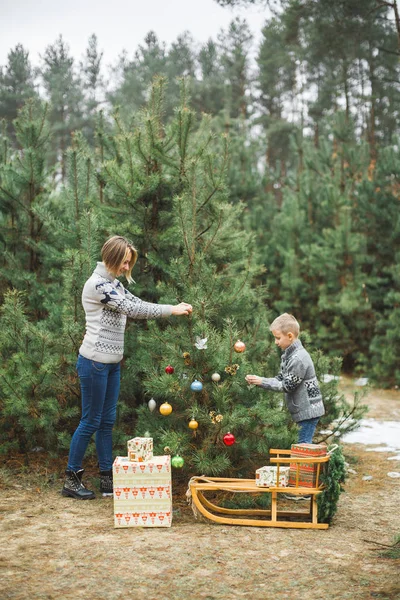 Bir anne ve oğlu, Noel ağacını açık havada, kış ormanında süsler. Yeni yıl süslemeleri, Noel çelenkli ahşap kızak, ağacın altında hediyeler. — Stok fotoğraf