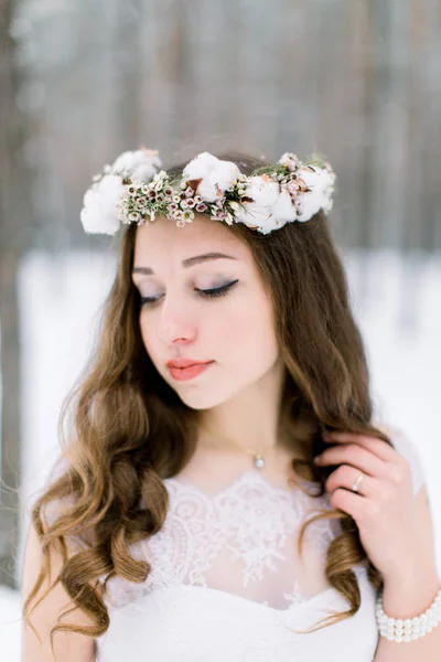 Hermosa novia tocando su pelo rizado, con corona floral de la banda de la cabeza, hecha de flores y algodón, de pie al aire libre, en el fondo del bosque de invierno —  Fotos de Stock