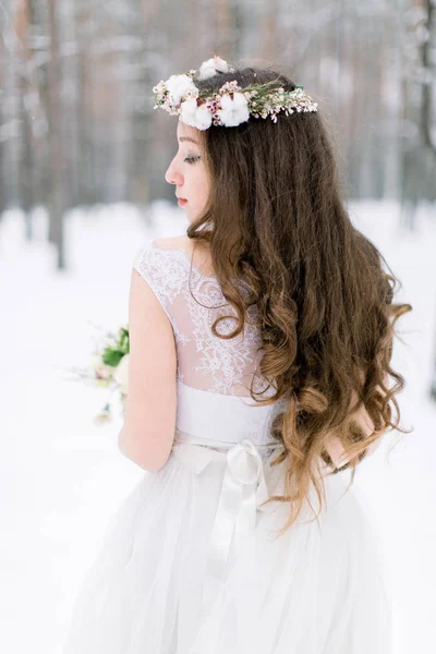 Mariage d'hiver, vue arrière de jolie mariée tenant bouquet debout dans la forêt d'hiver. Belle couronne de mariage d'hiver avec du coton et des fleurs sur la tête . — Photo