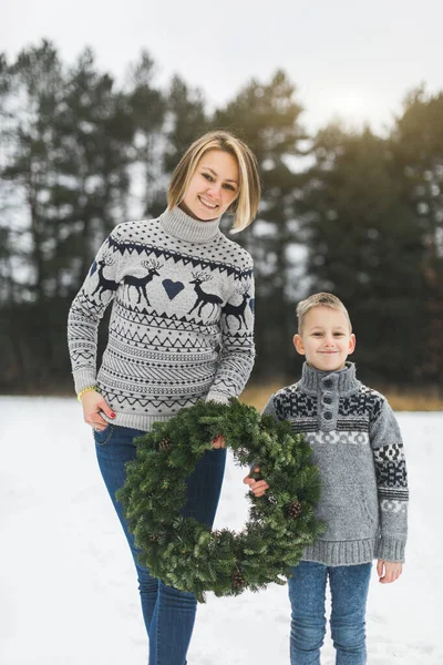 New Year and Christmas Holidays. Caucasian mom with her son holding decorated Christmas wreath in arms while standing outdoors on the background of winter snowy forest. — Stock Photo, Image