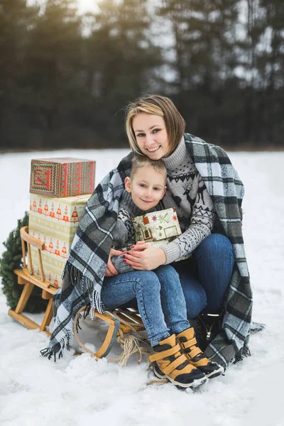 Mother with little son in winter park sledging. Mom and child sitting together on wooden decorated sledge with Christmas presents. Winter walk outdoors — Stock Photo, Image
