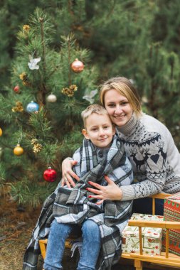 Anne ve küçük oğlu kış parkında kızak kayıyor. Anne ve çocuk, Noel hediyeleriyle süslenmiş ahşap bir kızakta birlikte oturuyorlar. Kış yürüyüşü, süslü Noel ağacı