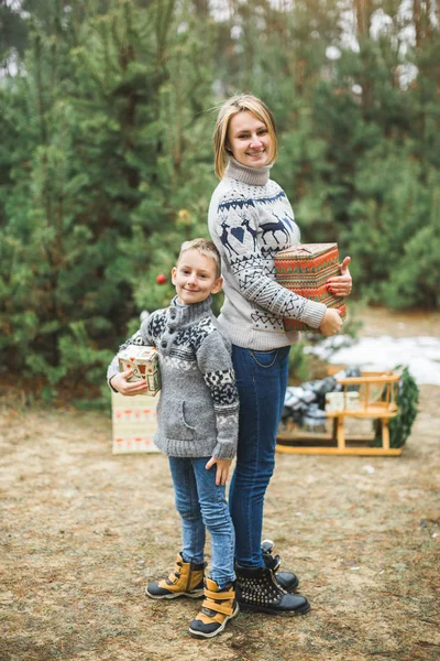 New Year and Christmas Holidays. Caucasian mom with her son holding decorated Christmas presents in arms while standing outdoors on the background of winter snowy forest. — Stock Photo, Image