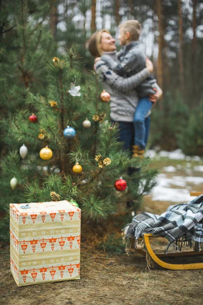 クリスマスプレゼントの箱、クリスマスプレゼントの木製のサンタのそり、冬の森と母と息子の抱擁の背景にクリスマスツリーを飾りました。メリークリスマスとハッピーニューイヤーコンセプト. — ストック写真